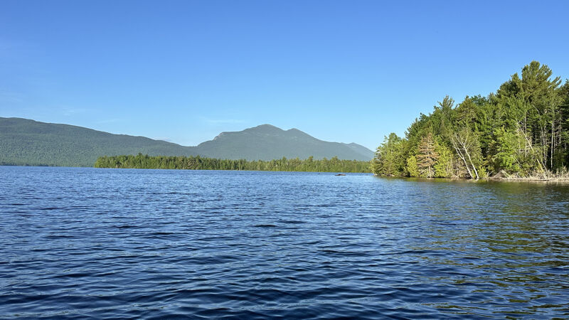 The image shows a serene lake scene under a clear blue sky. The water is calm, reflecting the sky and the distant mountains. Lush green trees line the shore, adding to the natural beauty of the landscape. The overall impression is one of peace and tranquility.